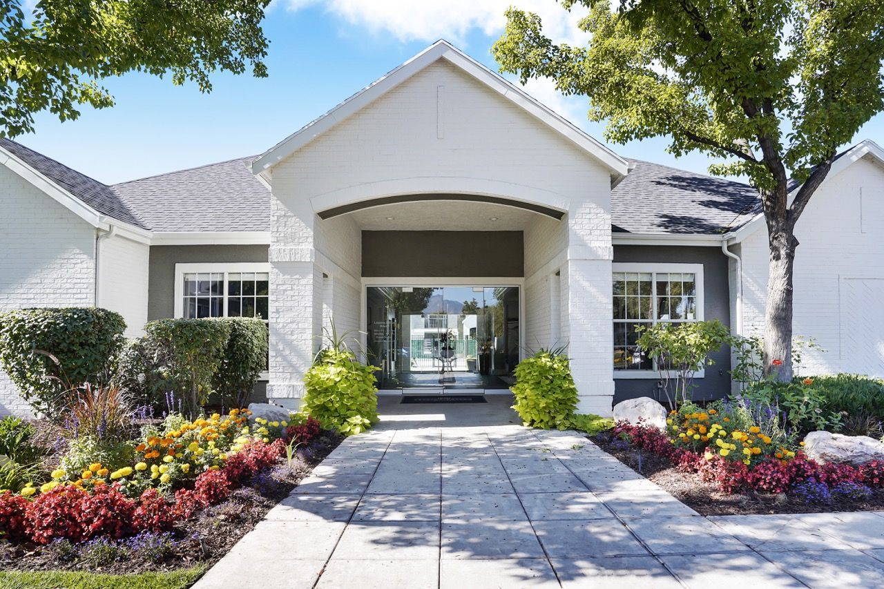 Front entrance of a white apartment community with landscaped flower beds and a walkway.