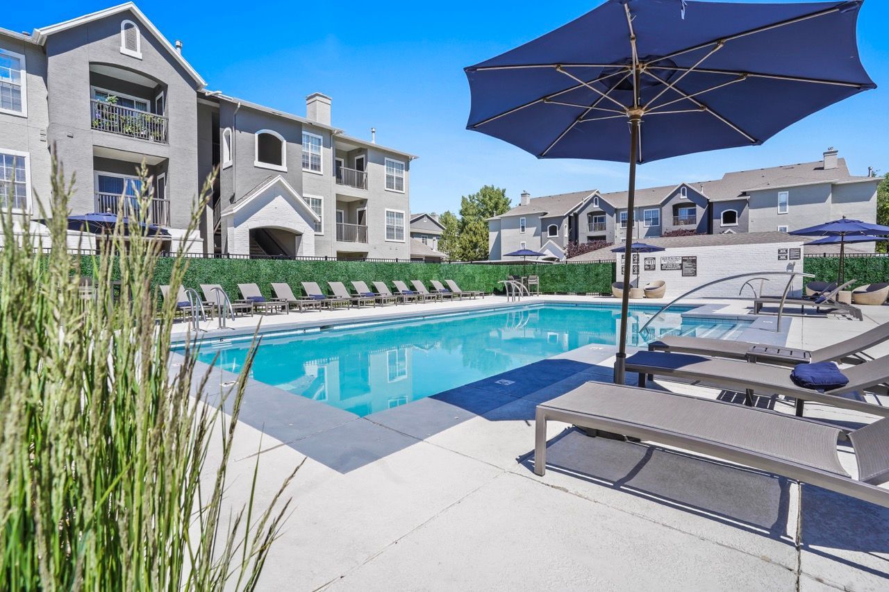 Outdoor pool area at a multi-building apartment community with lounge chairs and blue umbrellas.