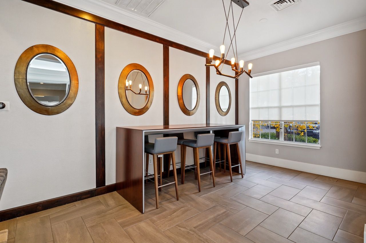 Interior community lounge with a high-top table, four stools, round mirrors on a paneled wall, and a chandelier.