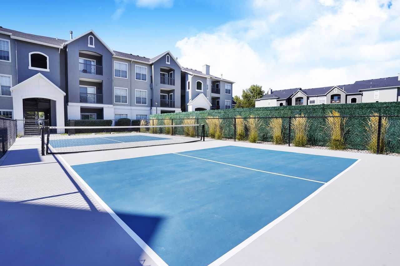 Outdoor community tennis court with a net, beside apartment buildings.