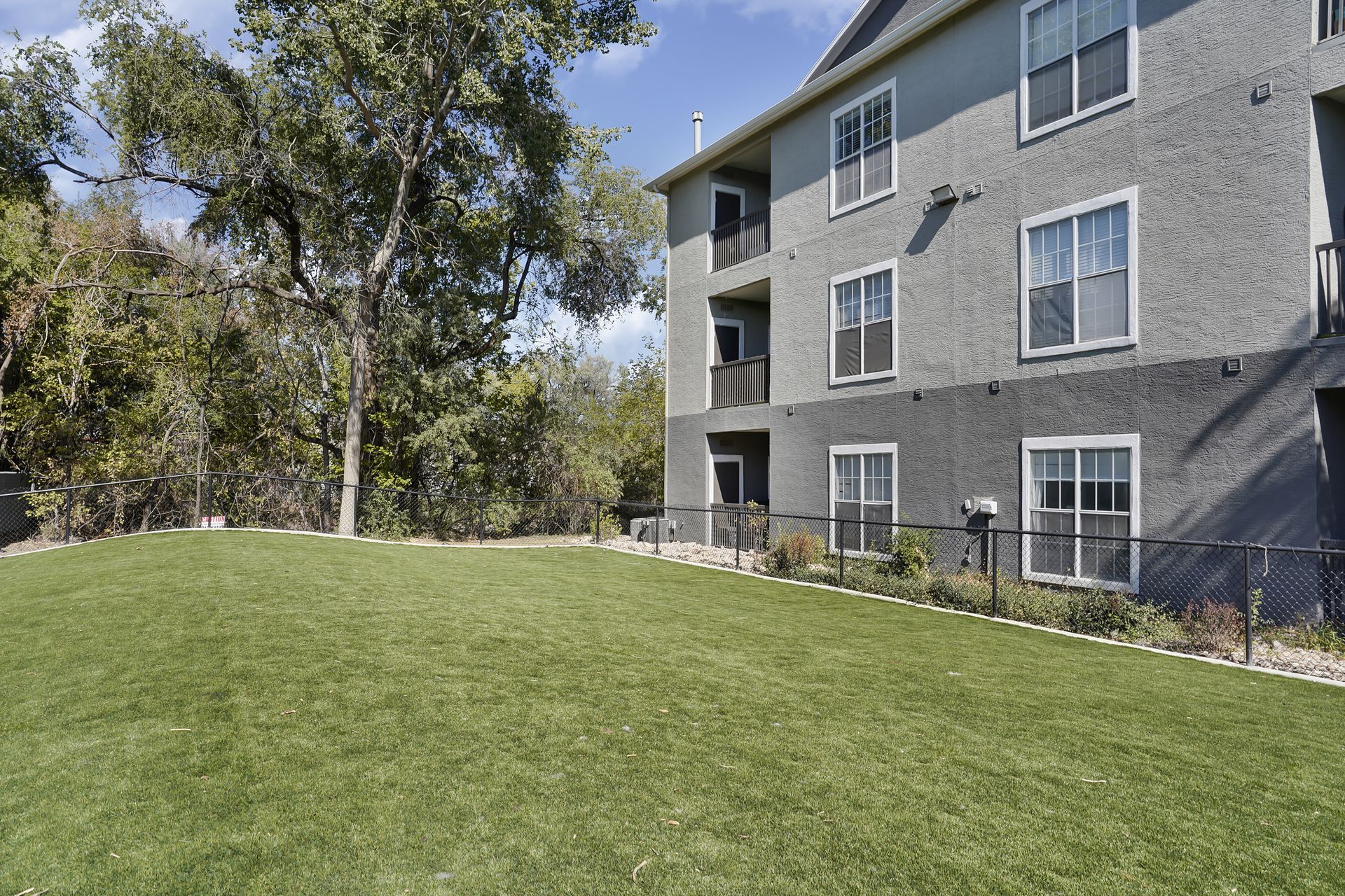 Green lawn next to a three-story building with dark gray exterior and windows. Trees in the background at Preston Hollow in Millcreek, UT.