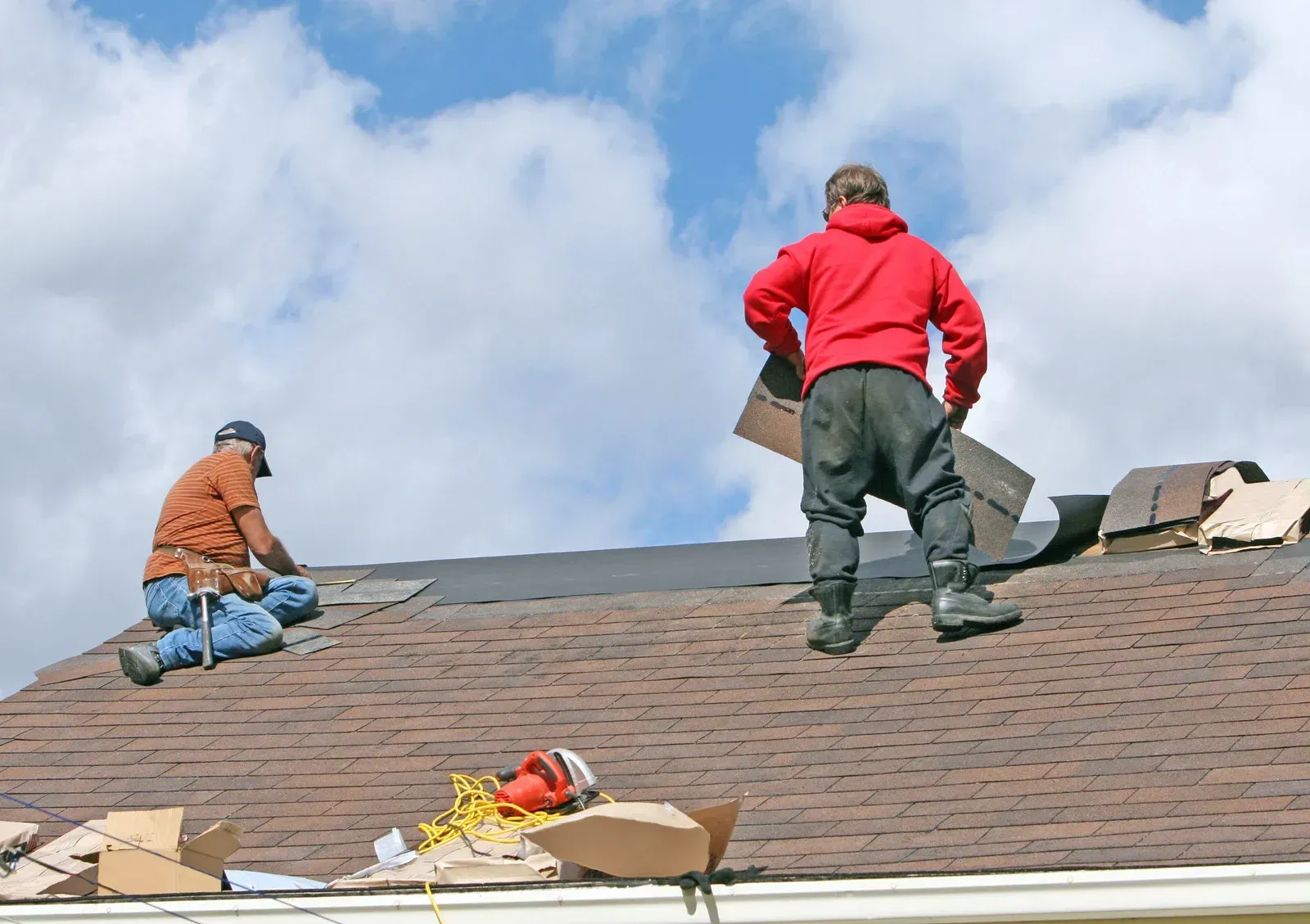 Two roofers repairing a shingled roof under a blue sky, with tools and supplies nearby