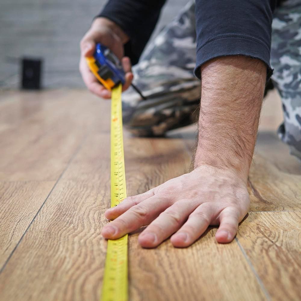 Person measuring a wooden floor with a yellow tape measure and one hand braced on the plank surface