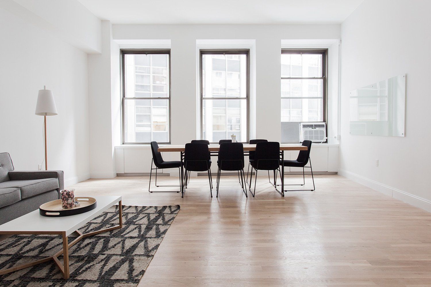 Bright minimalist dining room with black chairs, large windows, light wood floors, and a gray sofa