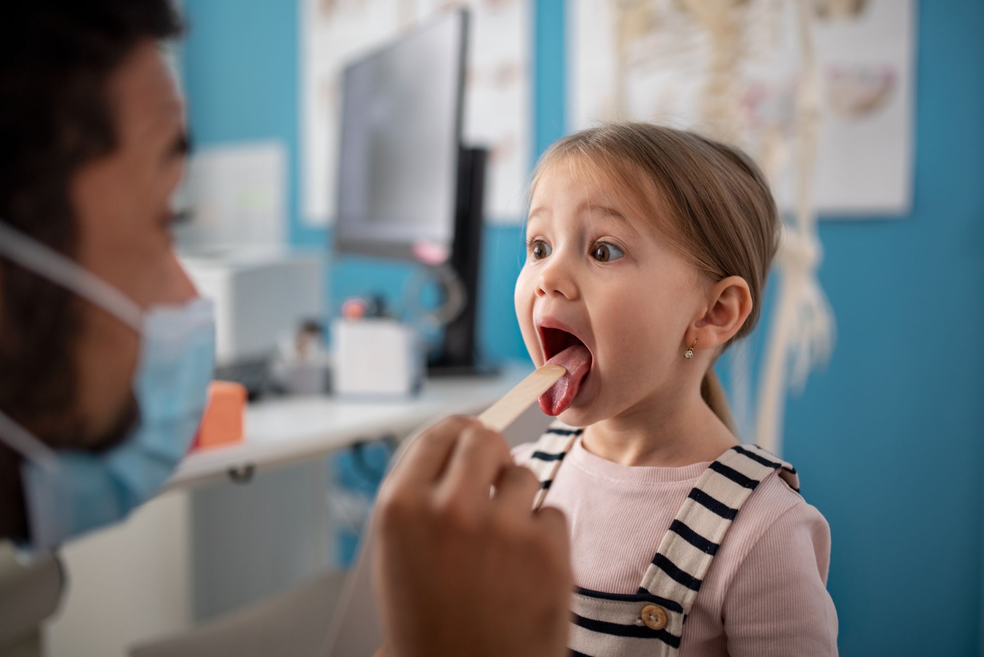 Child at doctor's office with mouth open for examination. Doctor with tongue depressor, wearing mask, looks on.