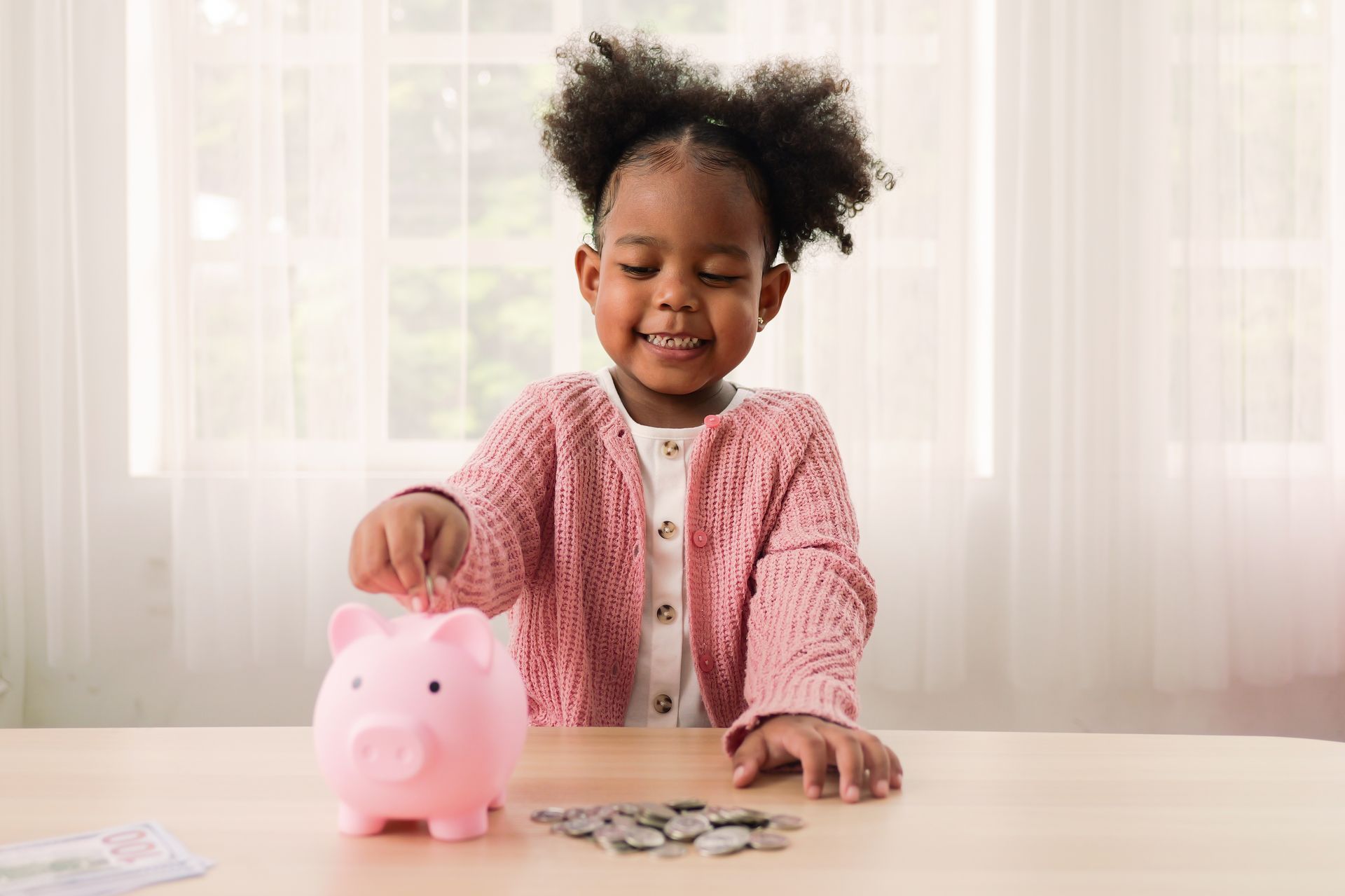 Girl in pink sweater putting coins in a pink piggy bank on a table.