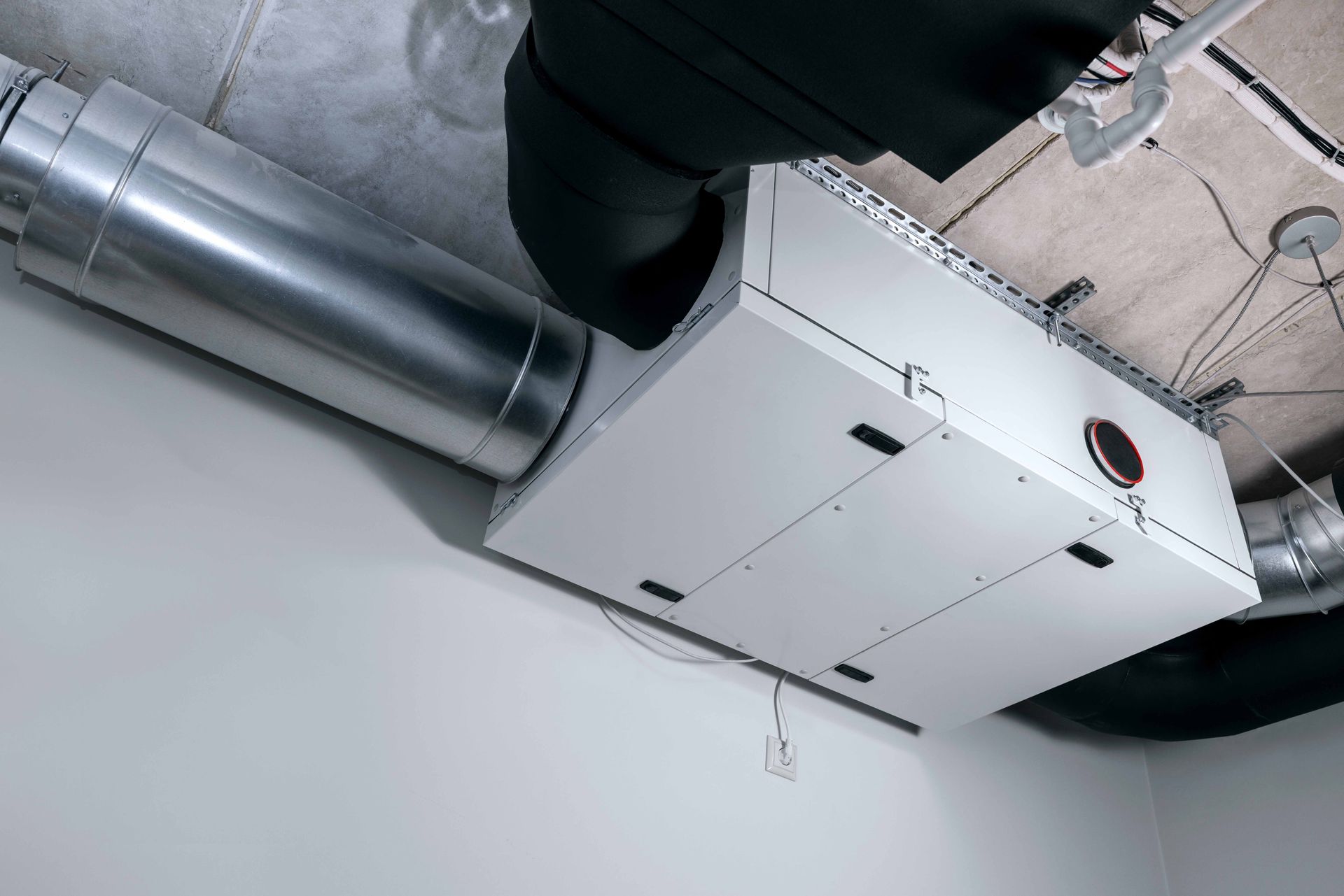 White air handler unit with ductwork in a room with a white wall and a grey ceiling.