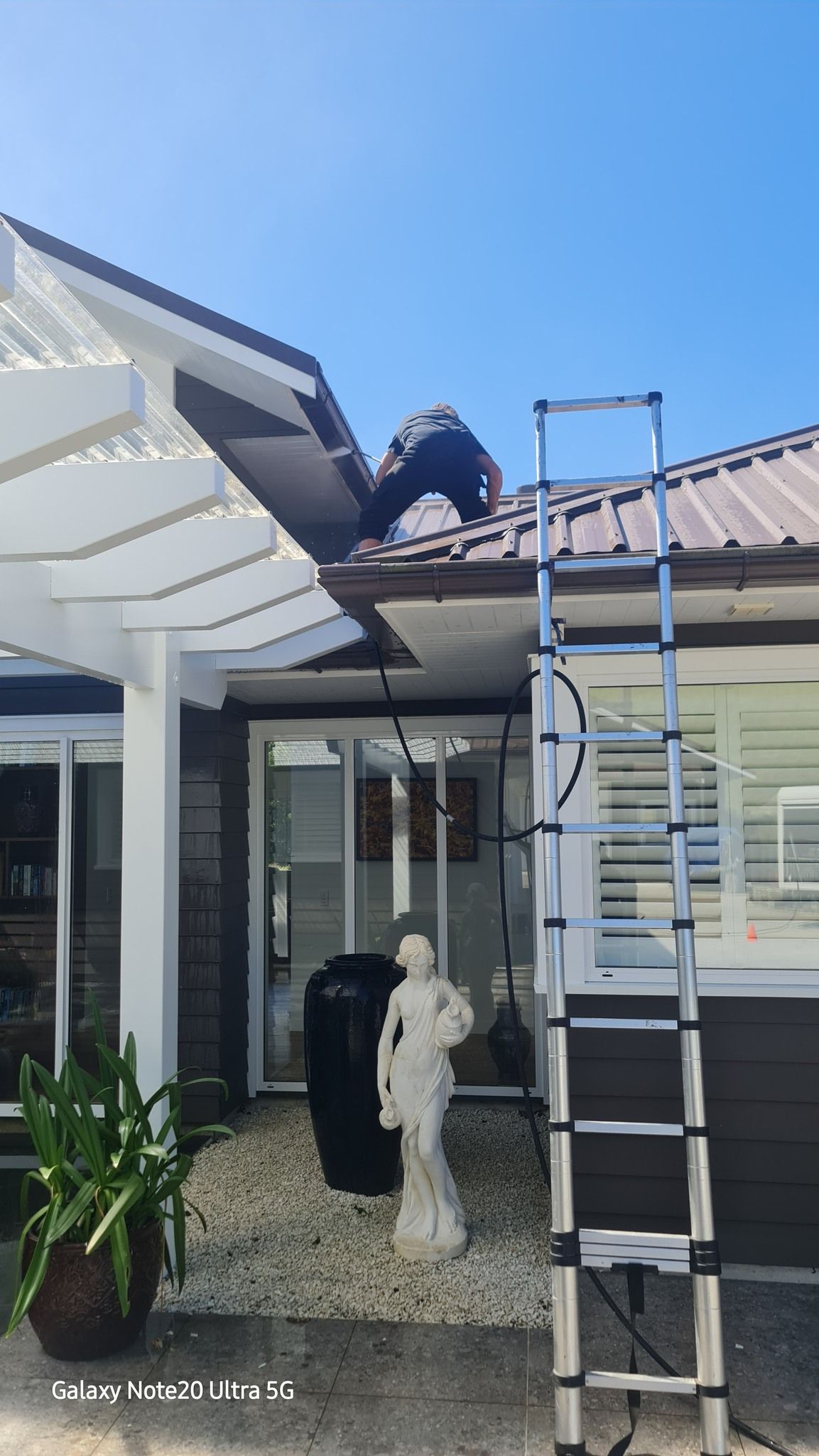A Man Is Standing on The Roof of A House Next to A Ladder - Auckland, NZ - Auckland Wash & Go Limited