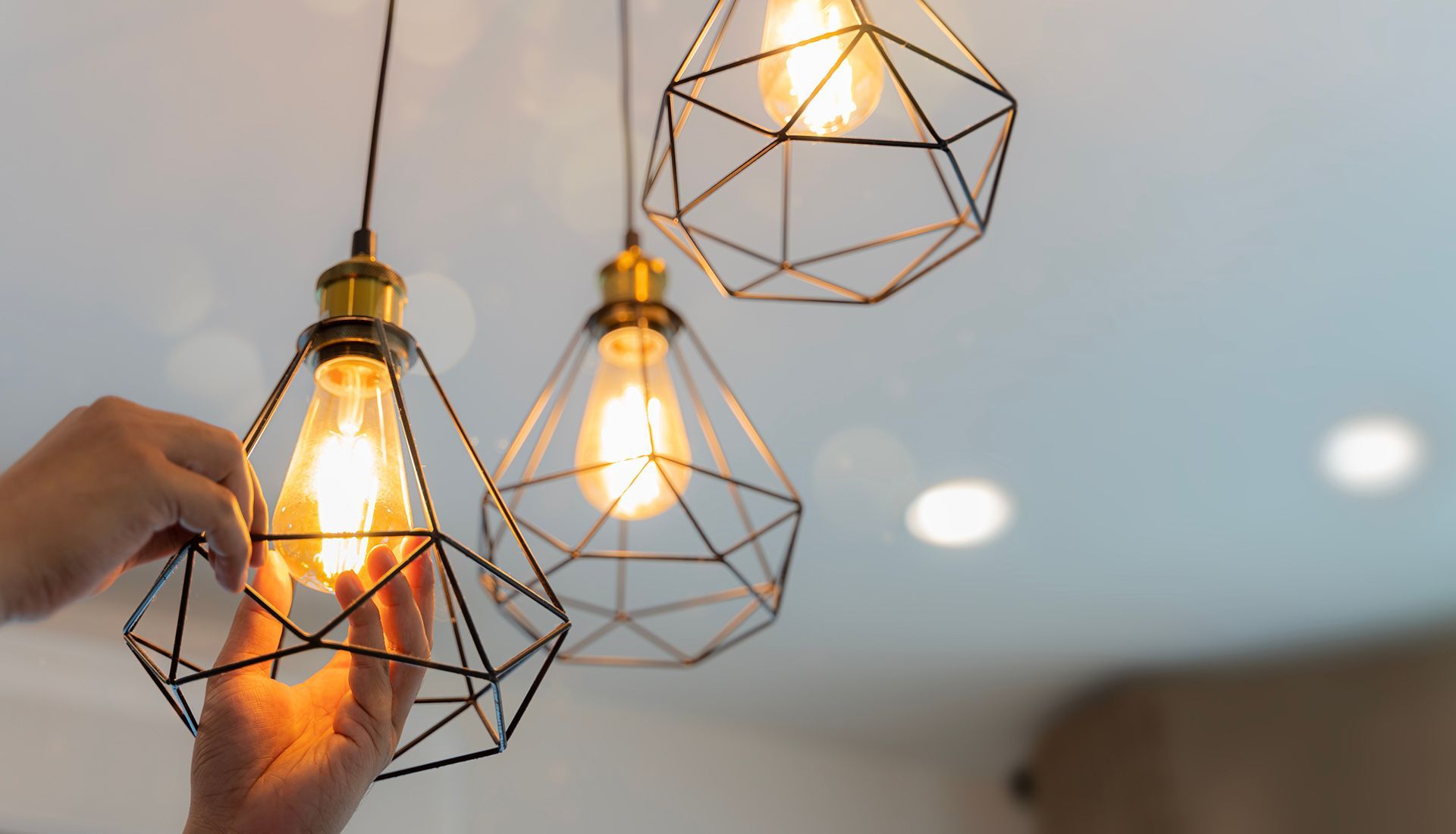 Person replacing a lightbulb in a geometric pendant lamp fixture, with two others hanging nearby.