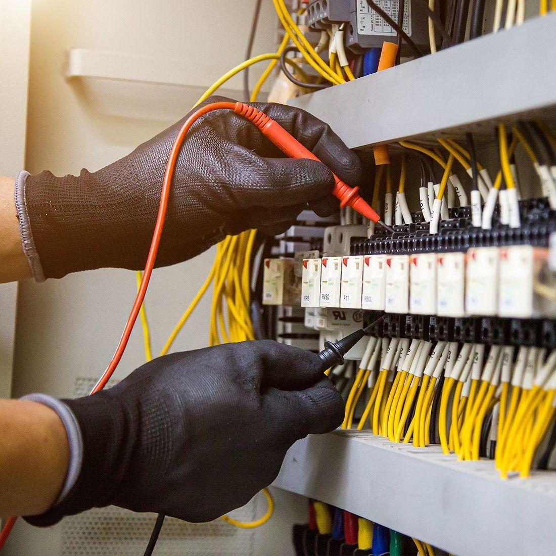 Hands wearing black gloves testing electrical wiring with a multimeter in a panel. Yellow and other colored wires visible.