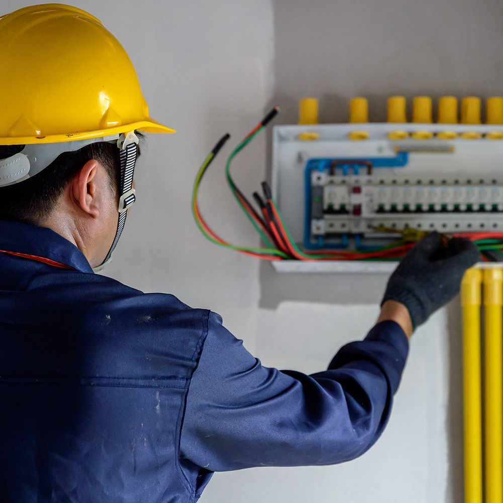 Electrician in a blue jumpsuit and yellow hard hat working on a circuit breaker panel, holding wires.