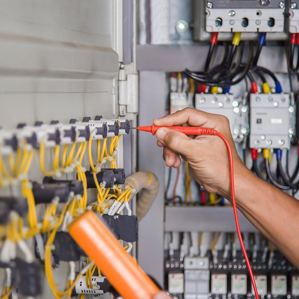 Person's hand testing electrical components with a red multimeter probe in an electrical panel filled with wires.