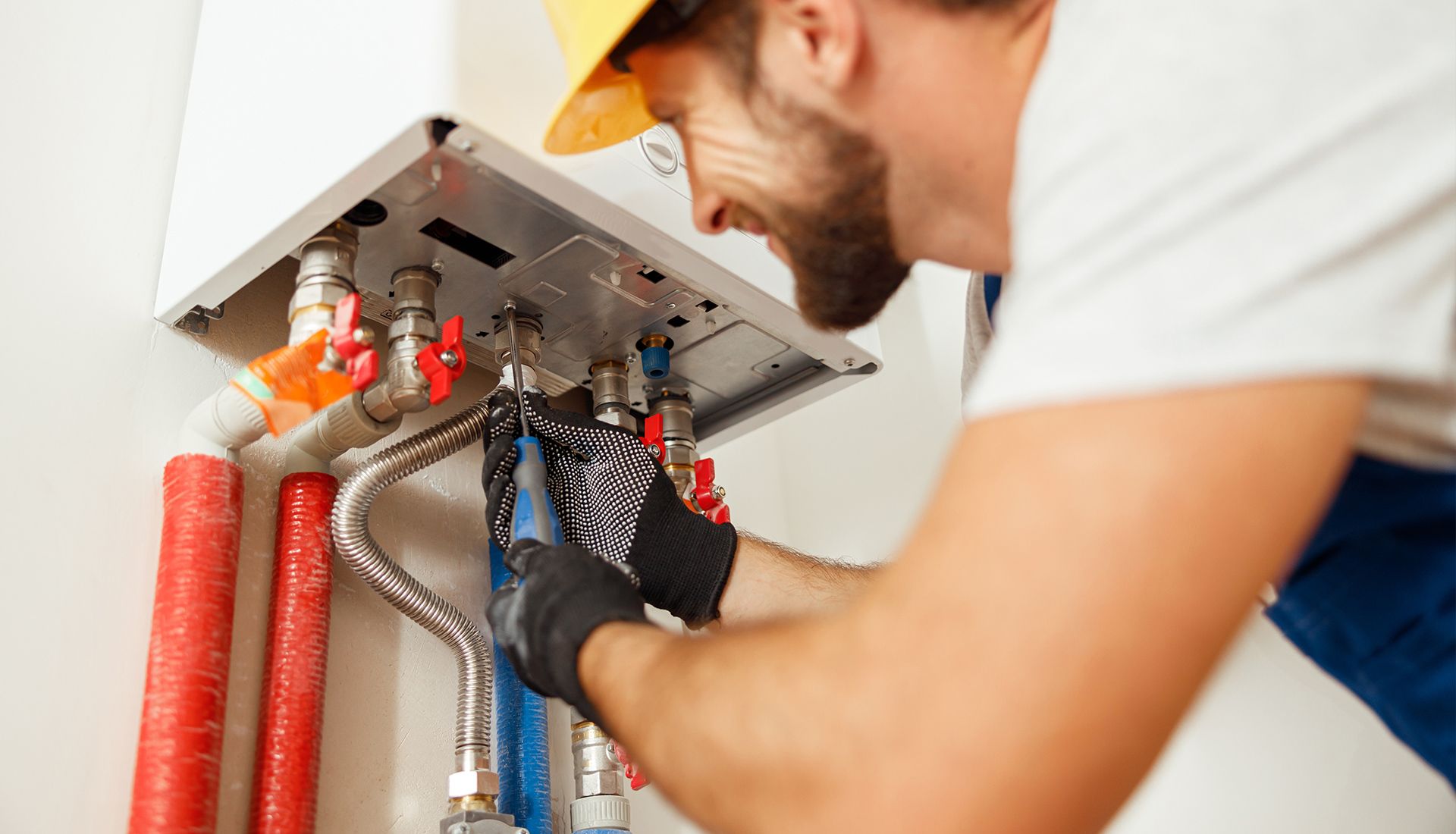 A smiling handyman in a hard hat and gloves works on a boiler with pipes, indoors.