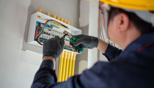 Electrician in blue uniform and hard hat working on an electrical panel, wires and conduits visible on a white wall.