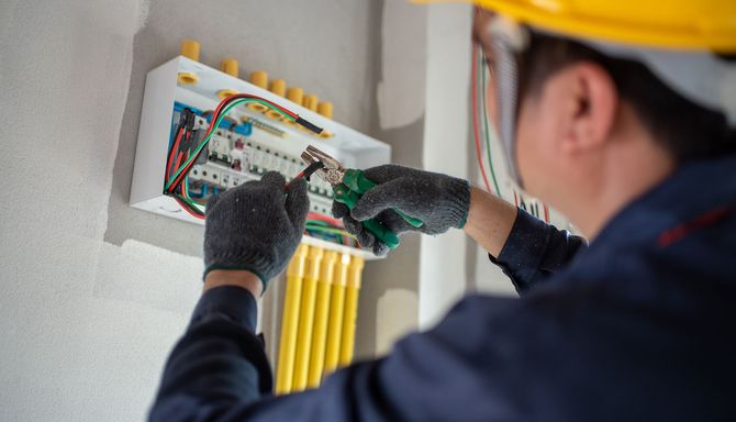 Electrician in blue uniform and hard hat working on an electrical panel, wires and conduits visible on a white wall.
