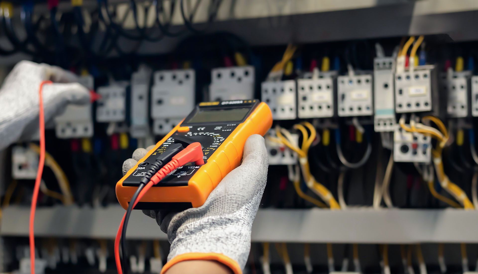 A person uses a multimeter to test electrical components in a control panel, wearing work gloves.
