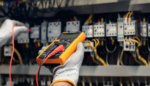A person uses a multimeter to test electrical components in a control panel, wearing work gloves.
