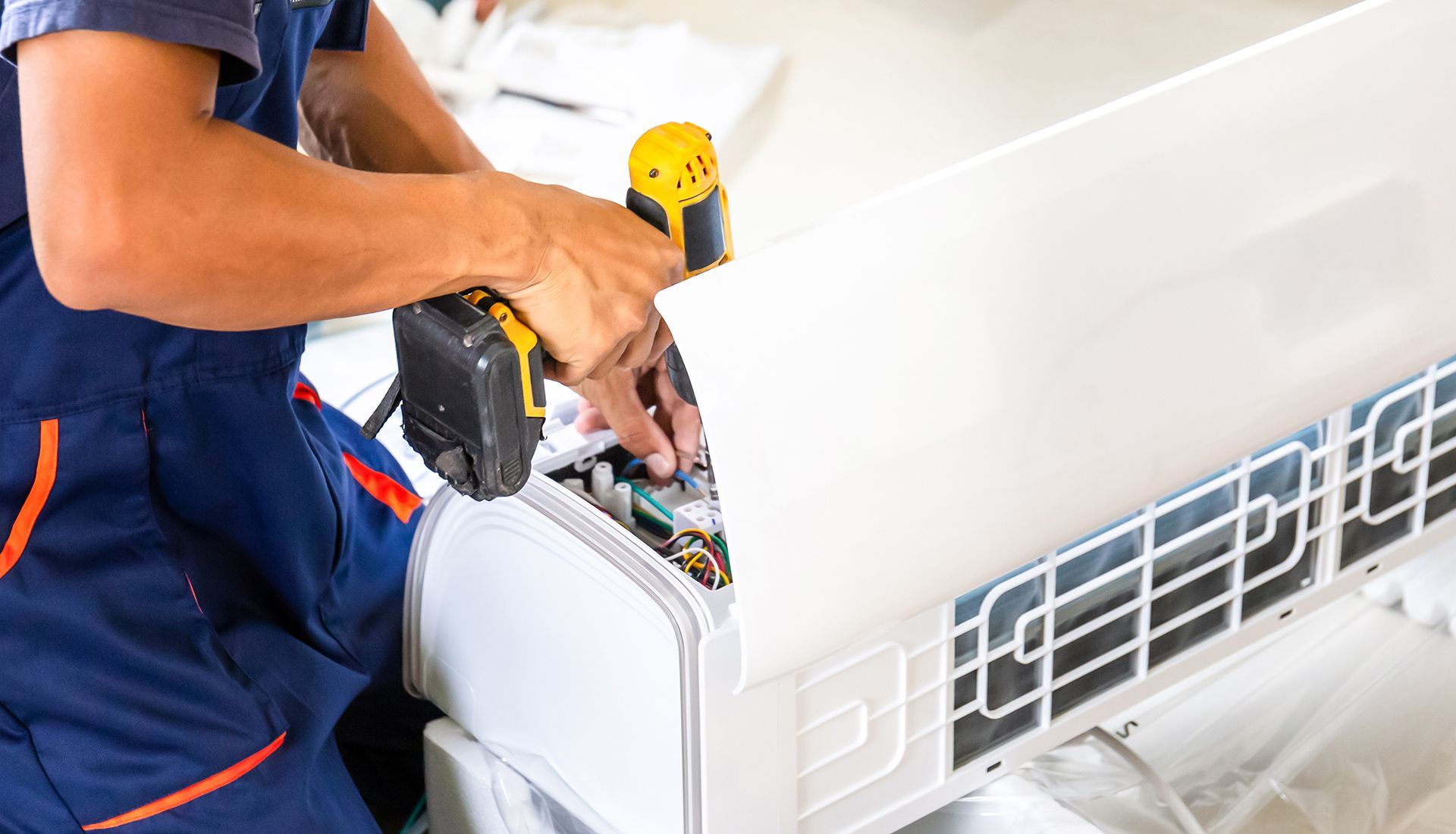 A person in blue overalls uses a power drill to install or repair a white air conditioning unit.