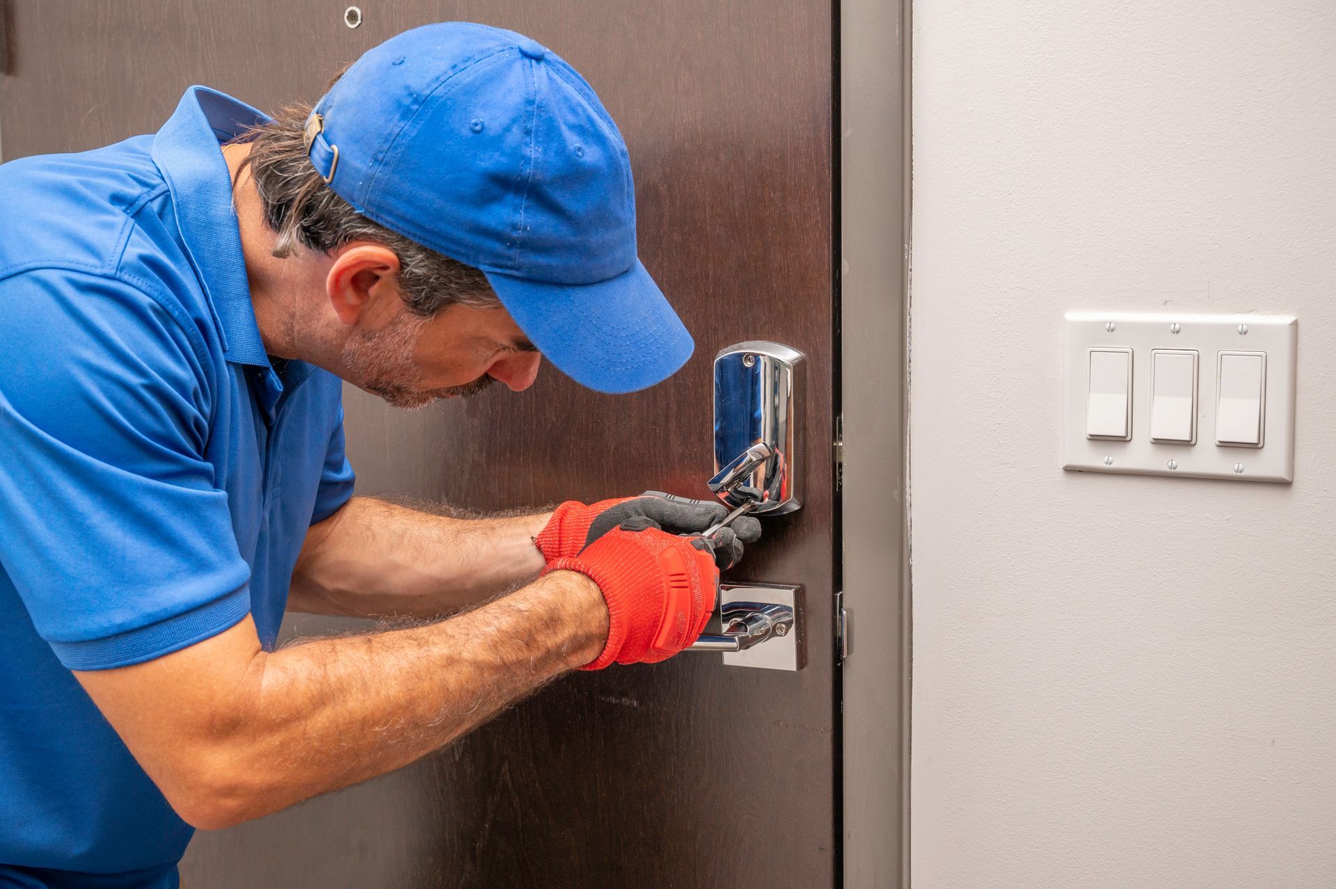 An emergency locksmith, in a blue uniform, is using a screwdriver on a deadbolt lock.