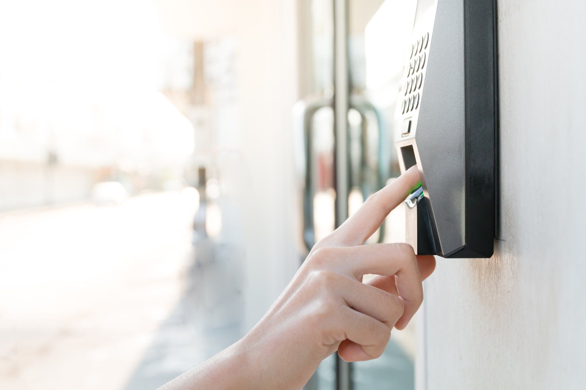 Person's finger touching a black biometric scanner mounted on a white wall near a glass door.