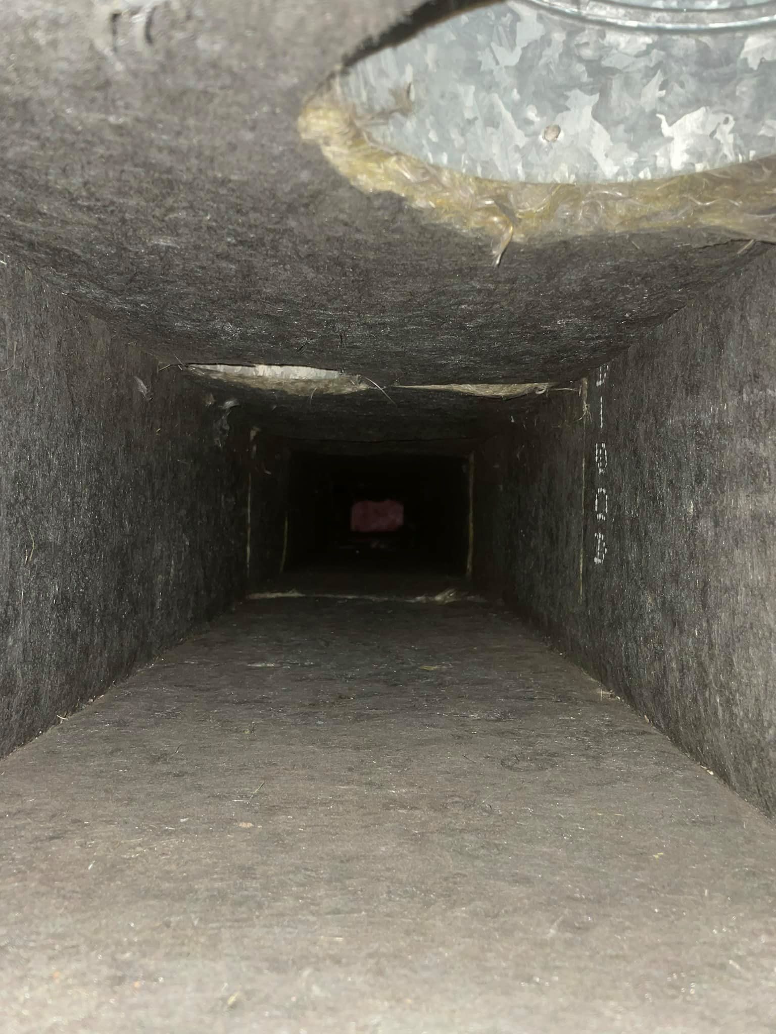 Dirty, rectangular air duct with dark walls, looking toward a dim opening.
