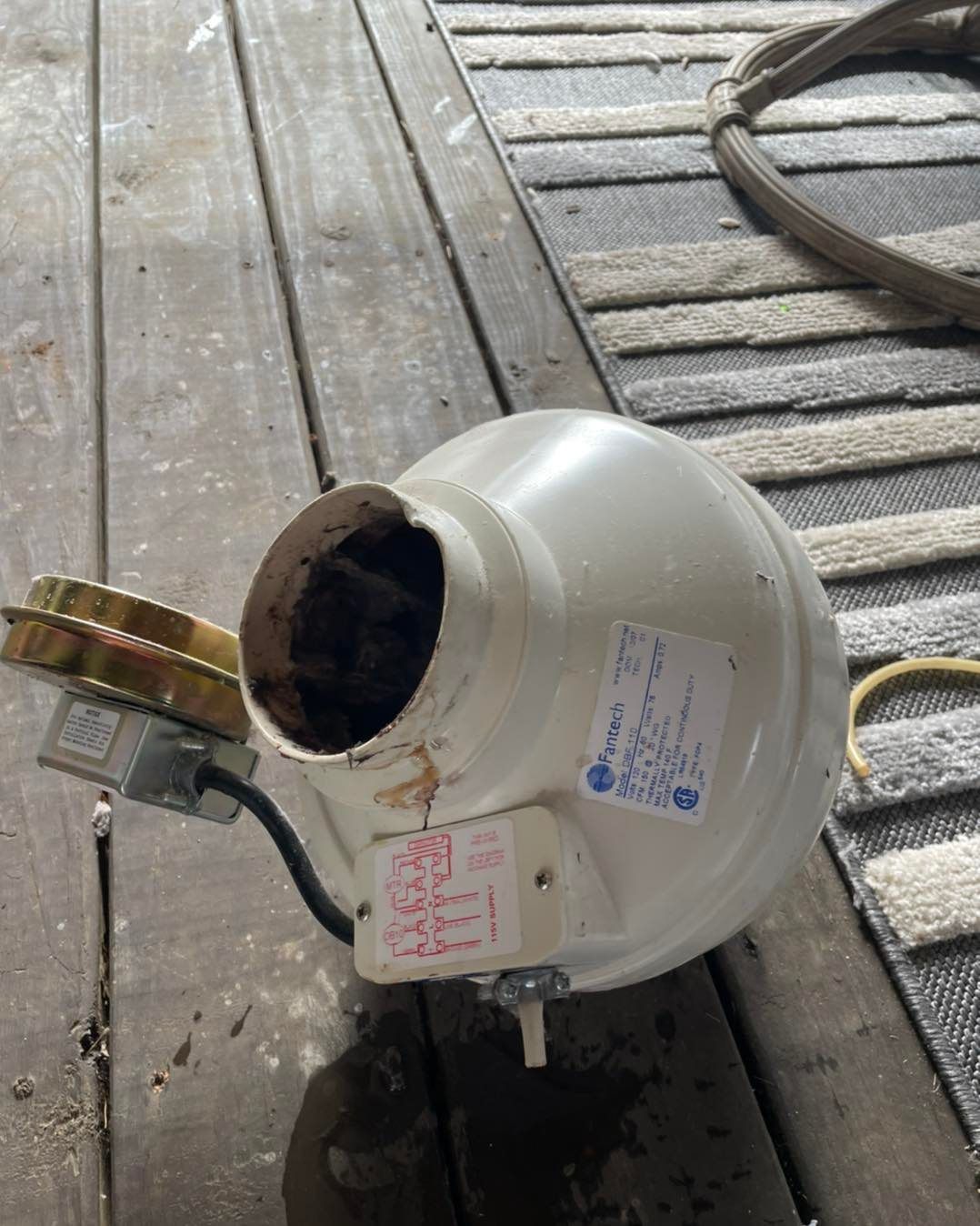 White, cylindrical inline duct fan with a dark interior on a wooden deck. Attached electrical components.