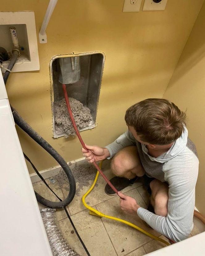 Person cleaning a laundry room vent. They are crouched, pulling a red cable from a wall cavity filled with debris.