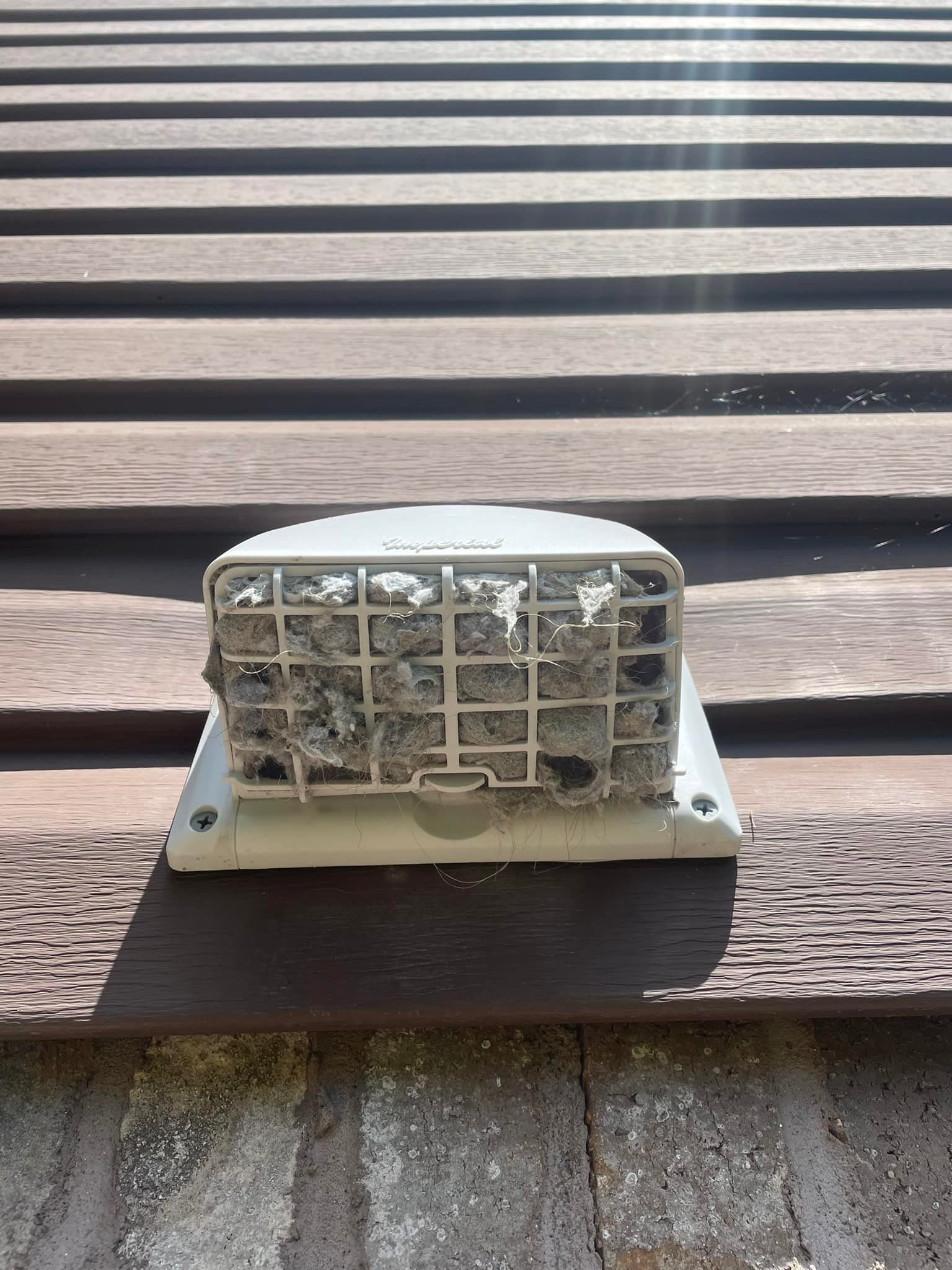 A white vent cover filled with debris is mounted on brown siding.