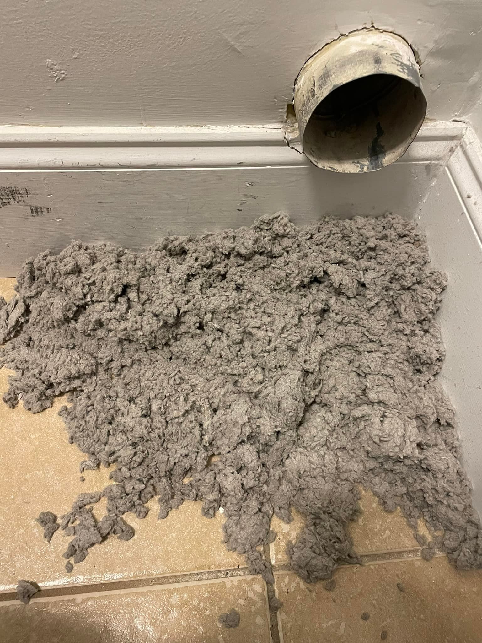 Pile of gray insulation on a tiled floor beneath a ventilation duct opening.
