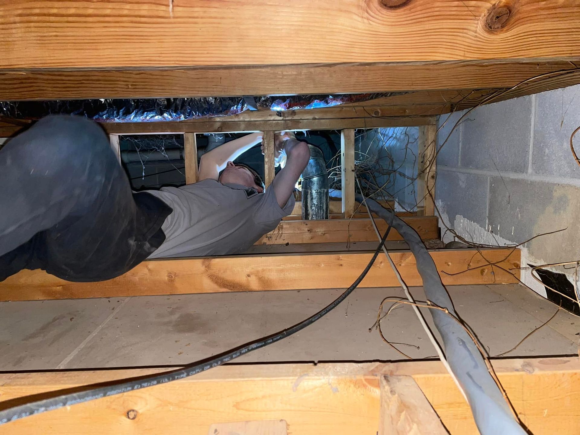 Man working in a crawl space, reaching toward pipes, surrounded by wood, wires, and insulation.