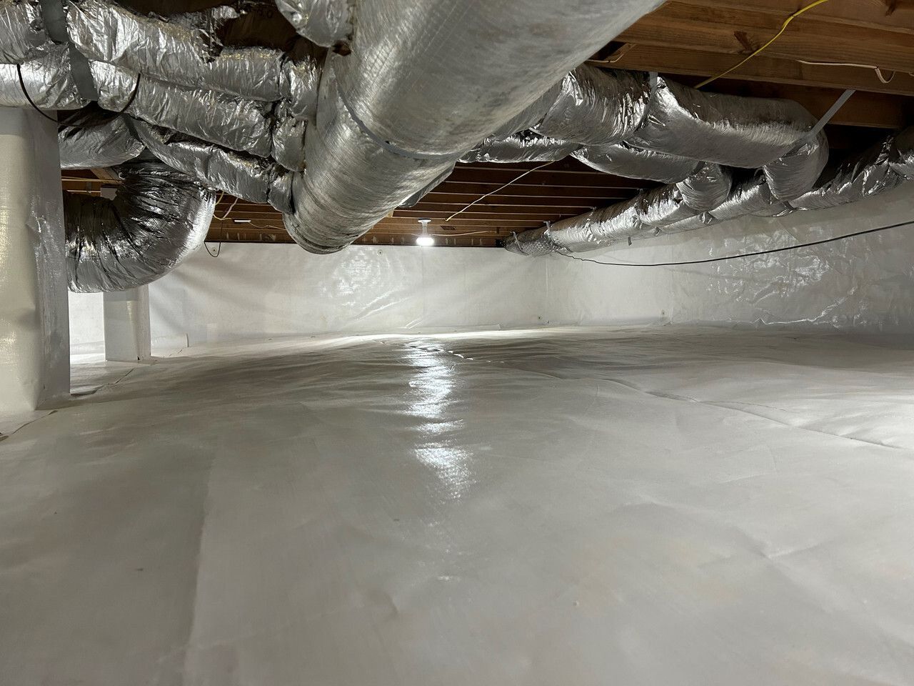 Crawl space with white vapor barrier, reflective ductwork, and exposed wooden joists.