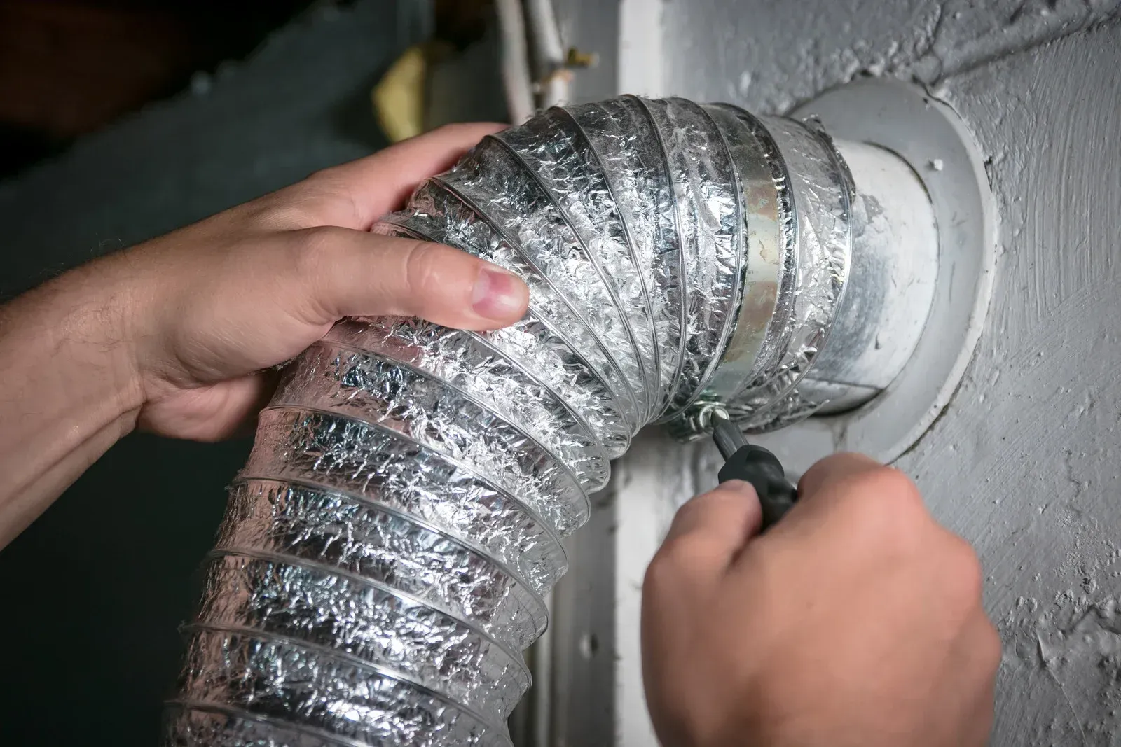 Hands attaching a flexible dryer vent hose to a wall outlet.