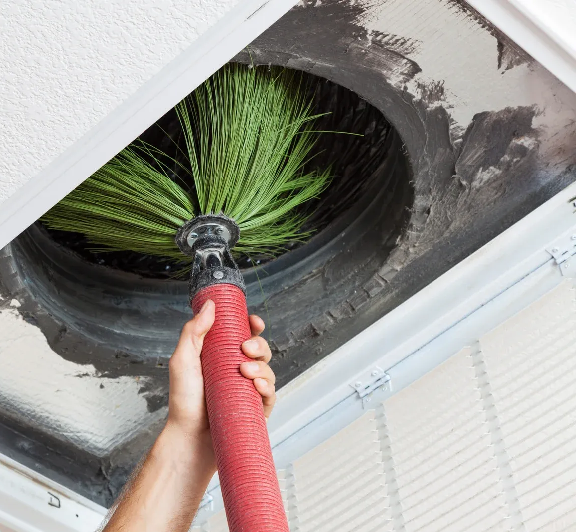 Person cleans HVAC duct with a brush. Red handle, green bristles, gray duct.