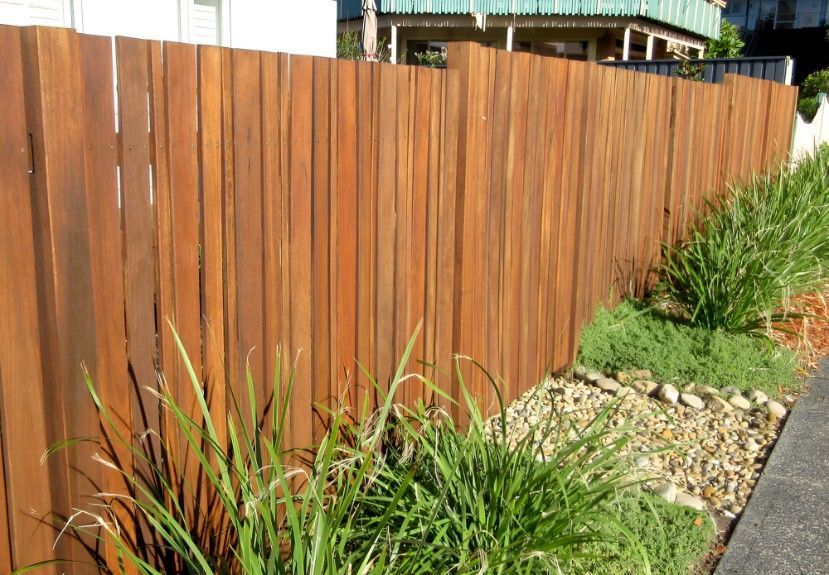 A Wooden Fence is Surrounded by Grass and Rocks Next to a Sidewalk — Harbour Fencing In Woolgoolga, NSW