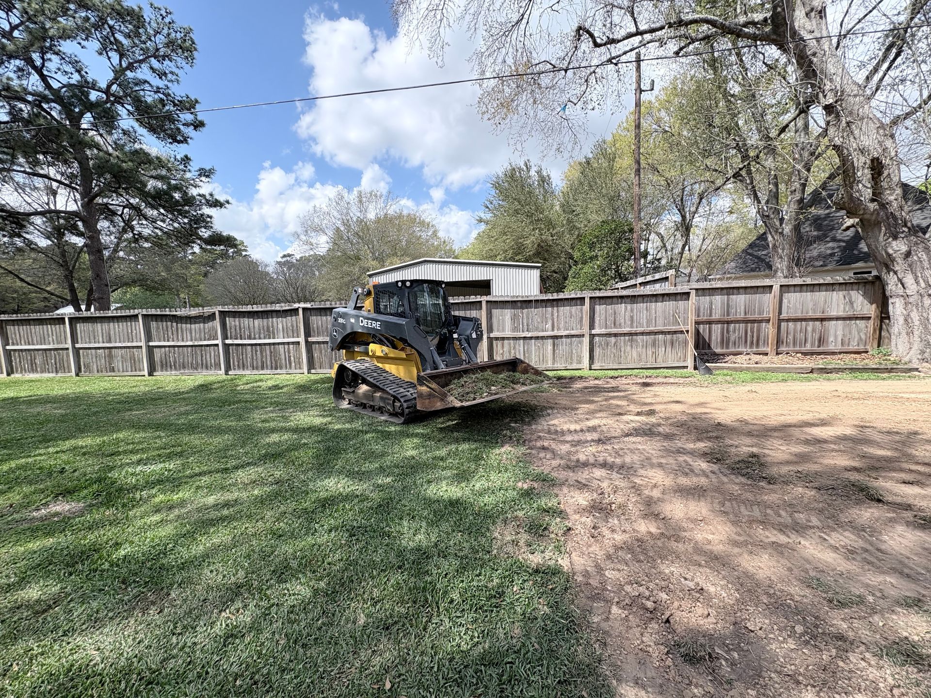 A yellow skid steer loader sits on a grassy lot next to a cleared dirt area in front of a wooden fence.