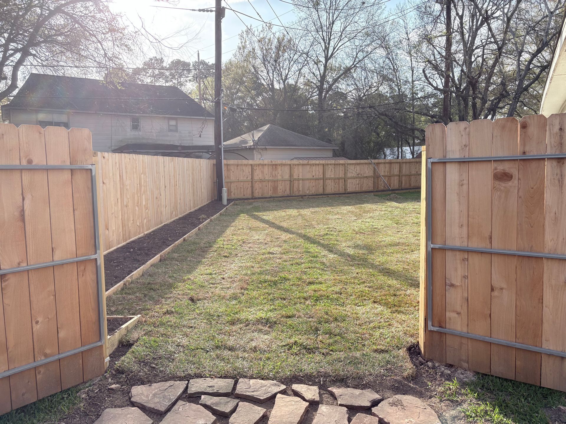 A yard viewed through an open wooden gate, featuring a stone path, grass lawn, and a surrounding wooden fence.