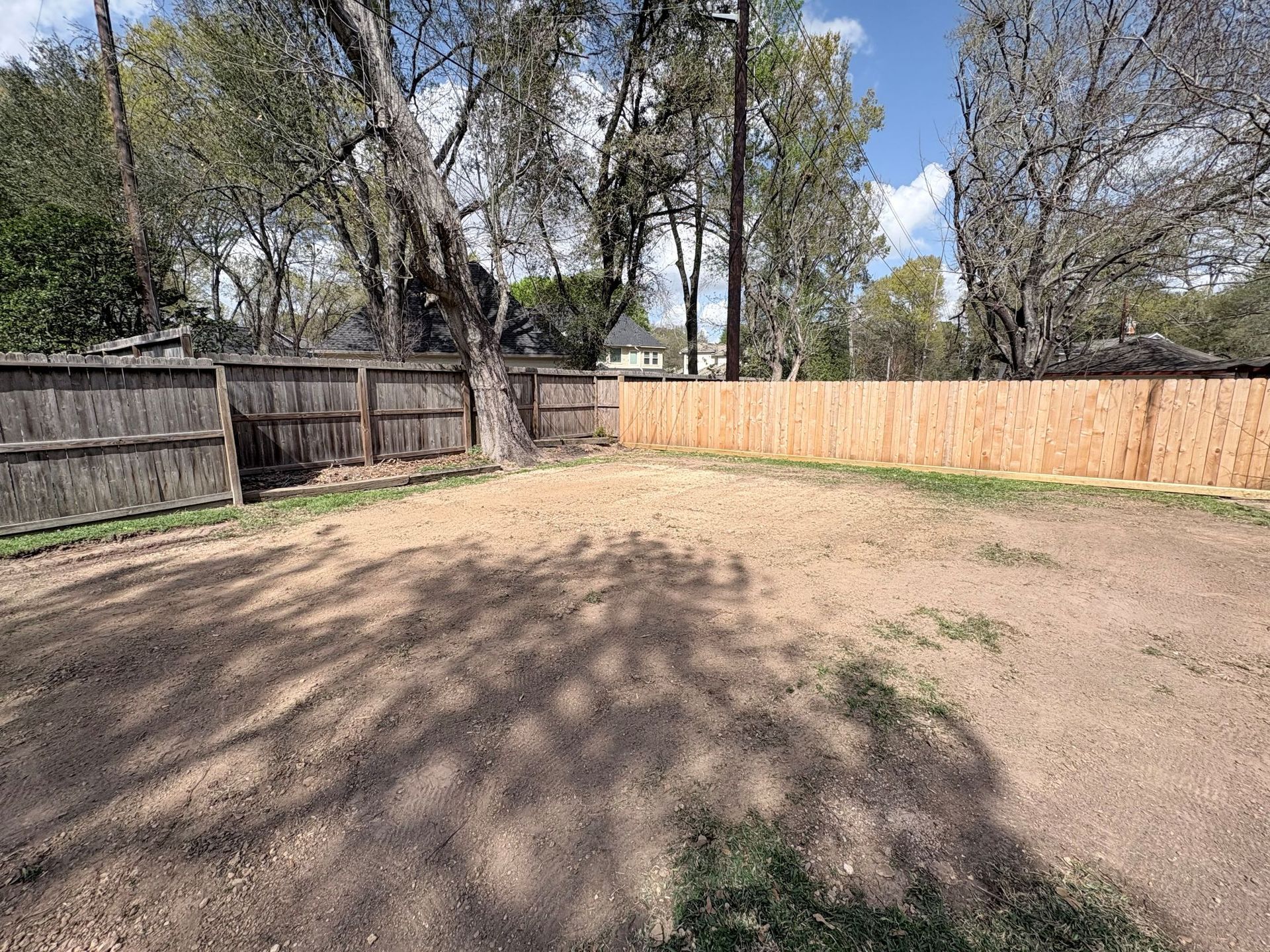 A fenced-in backyard with bare dirt ground, surrounded by mature trees under a clear blue sky.