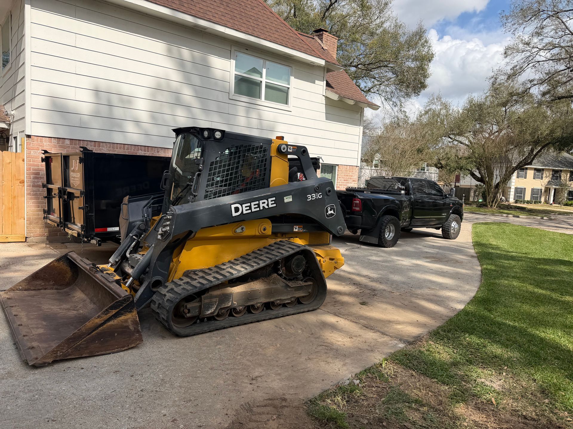 A yellow and black John Deere skid steer loader parked on a residential driveway next to a truck and dumpster.