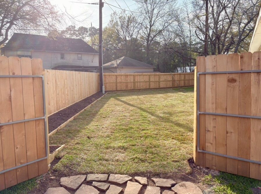 A stone path leads through an open wooden gate into a grassy backyard enclosed by a new wooden fence.