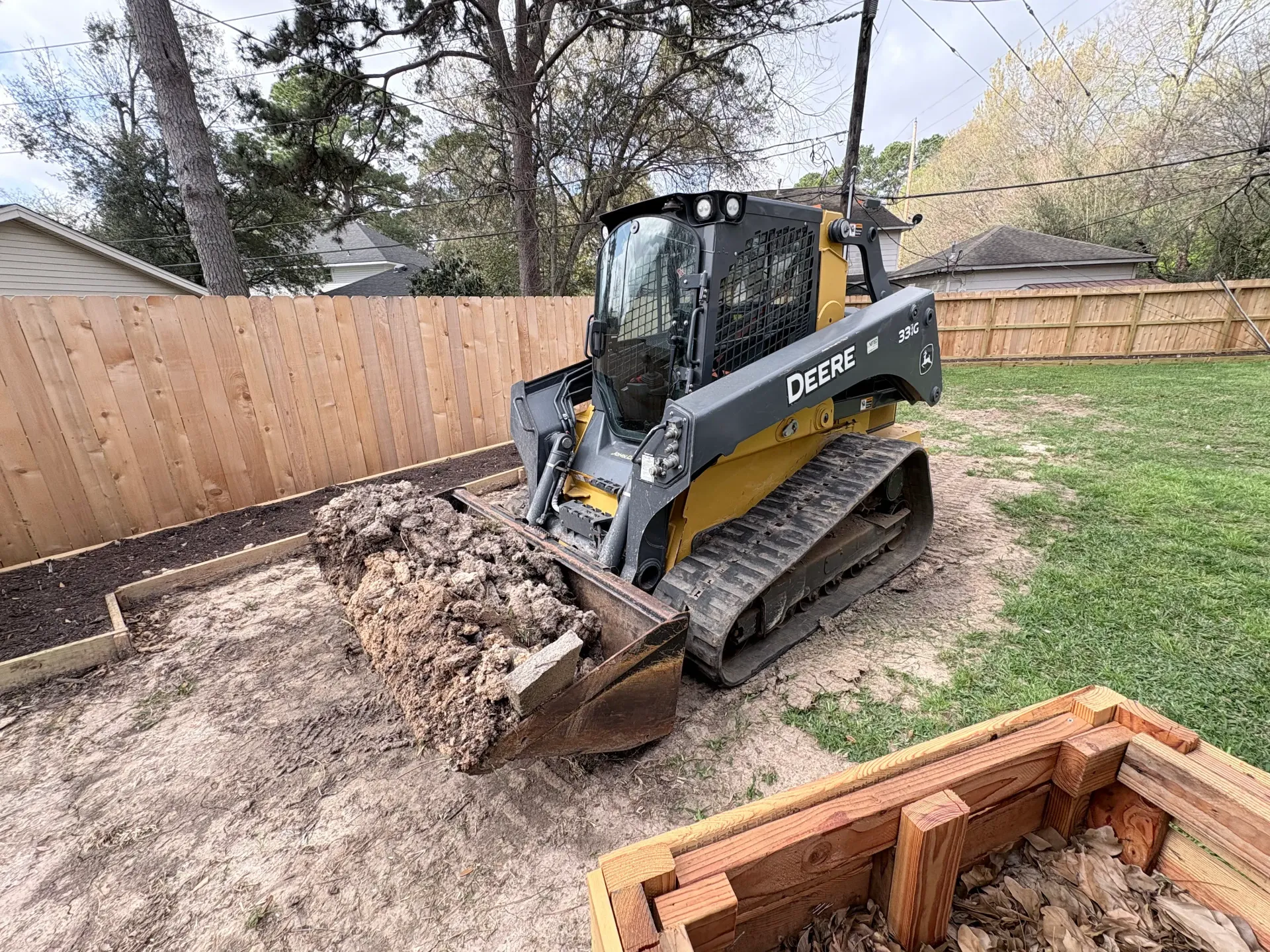 A yellow John Deere compact track loader scoops debris in a backyard next to a wooden fence and raised garden bed.