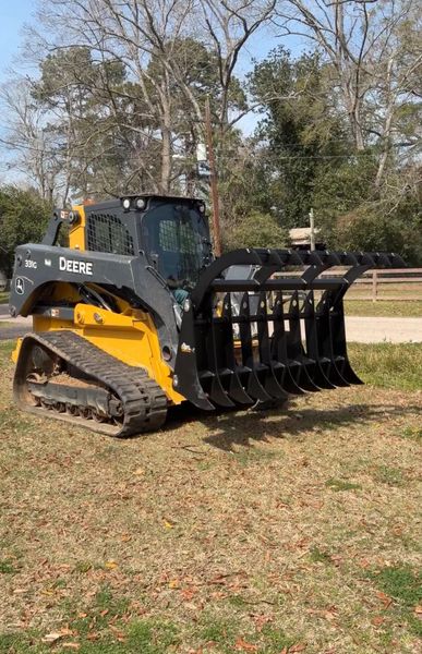 A yellow and black John Deere tracked skid steer loader equipped with a large metal root rake grapple on a grassy lot.