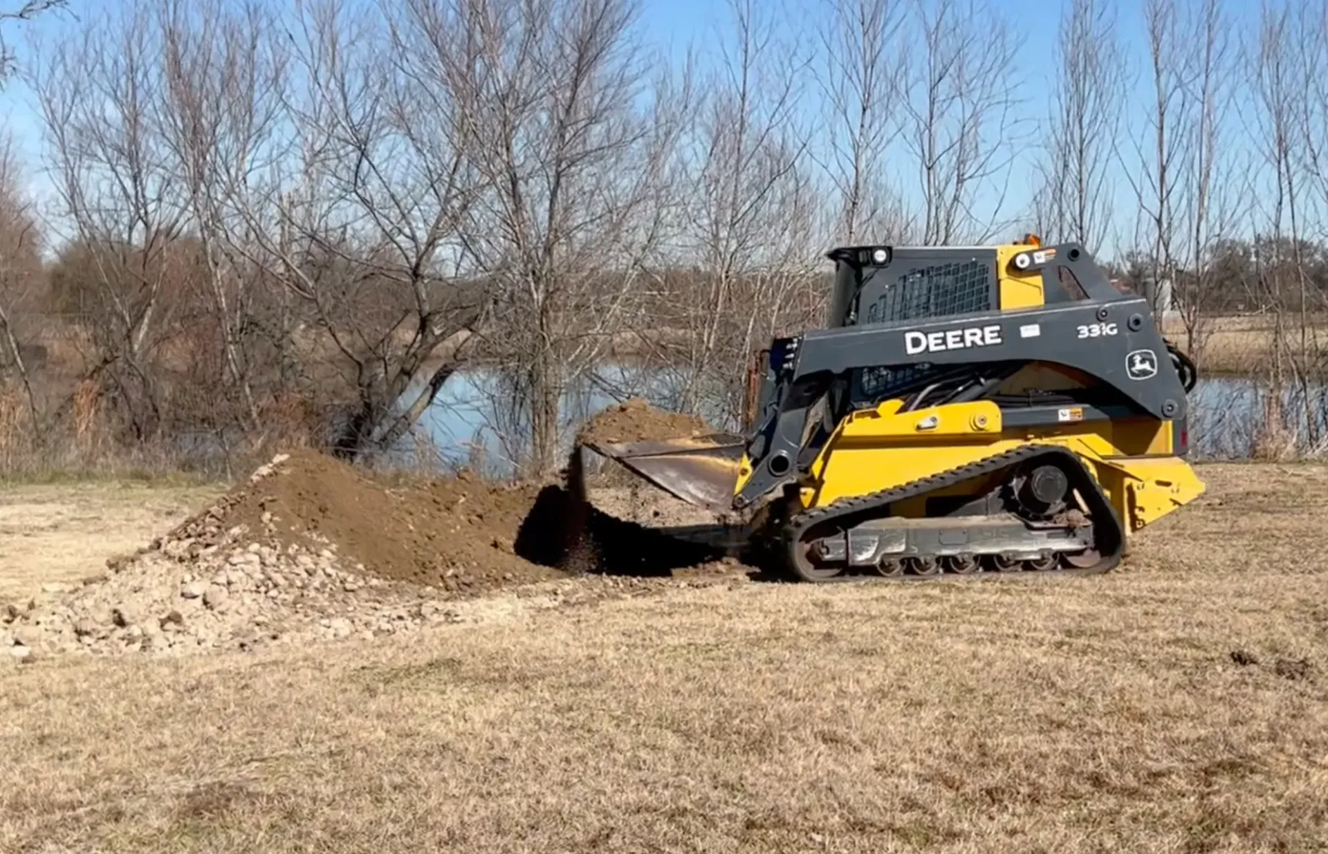 A yellow John Deere compact track loader pushes a mound of dirt in an outdoor field near a pond on a sunny day.