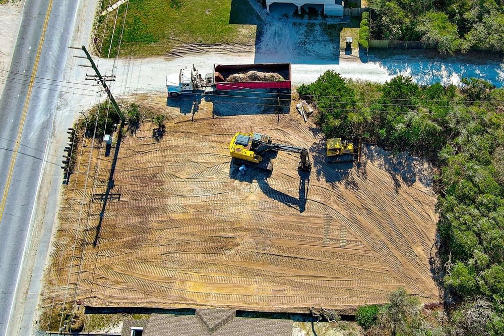 An aerial view of a construction site with heavy machinery, a red dump truck, and a cleared dirt lot next to a road.