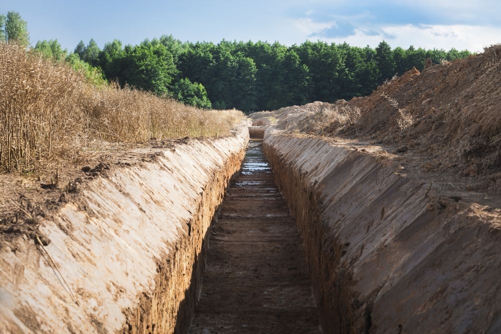 A long, deep trench dug into sandy soil, stretching toward a line of trees in the background under a blue sky.
