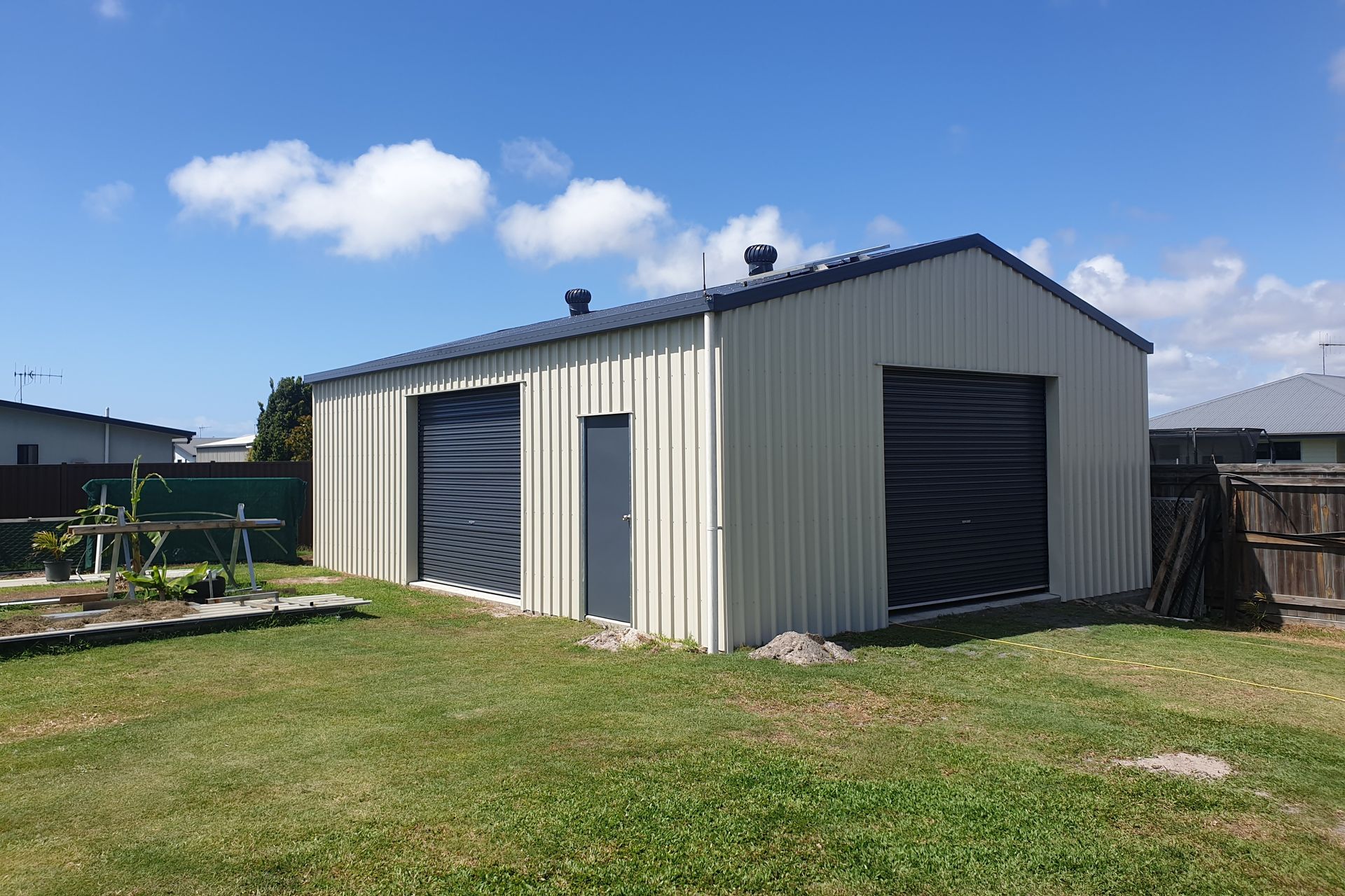 Tan metal garage with two bays and a small door on green lawn under a blue sky — Steel Homes Australia in Branyan, QLD
