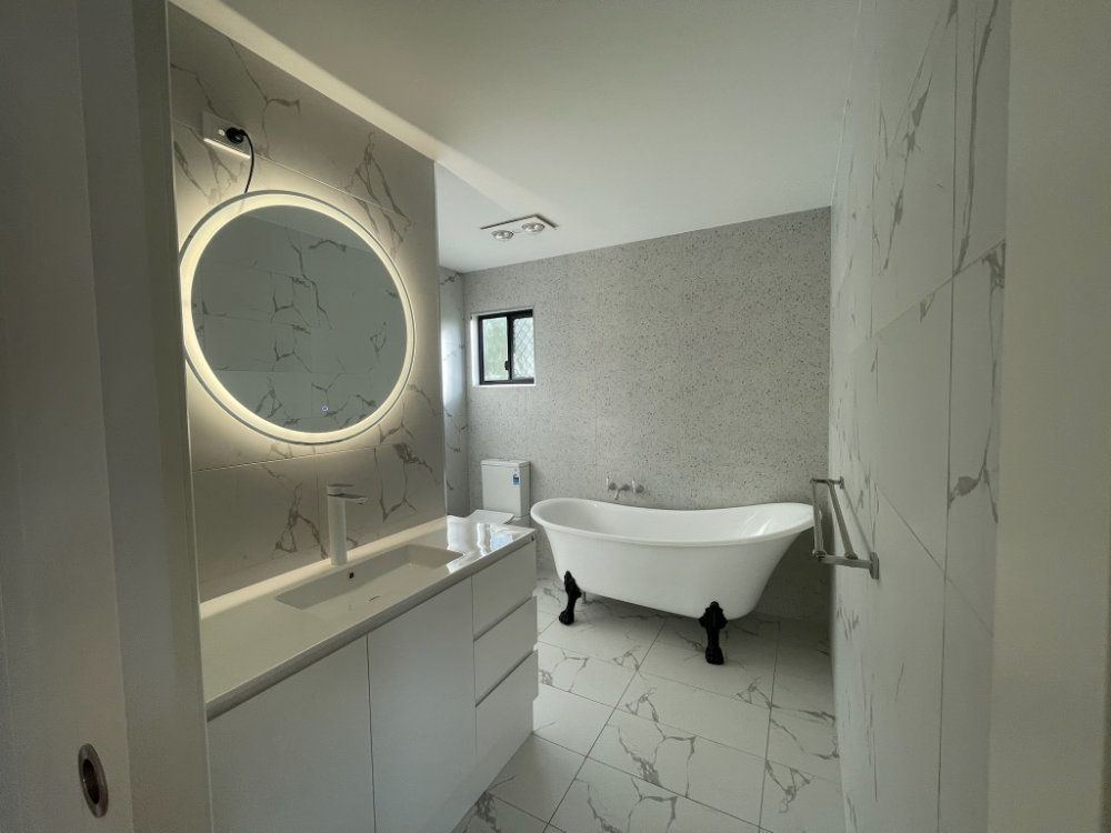 Modern bathroom with marble-look walls, a freestanding tub, and a circular lit mirror — Steel Homes Australia in Branyan, QLD