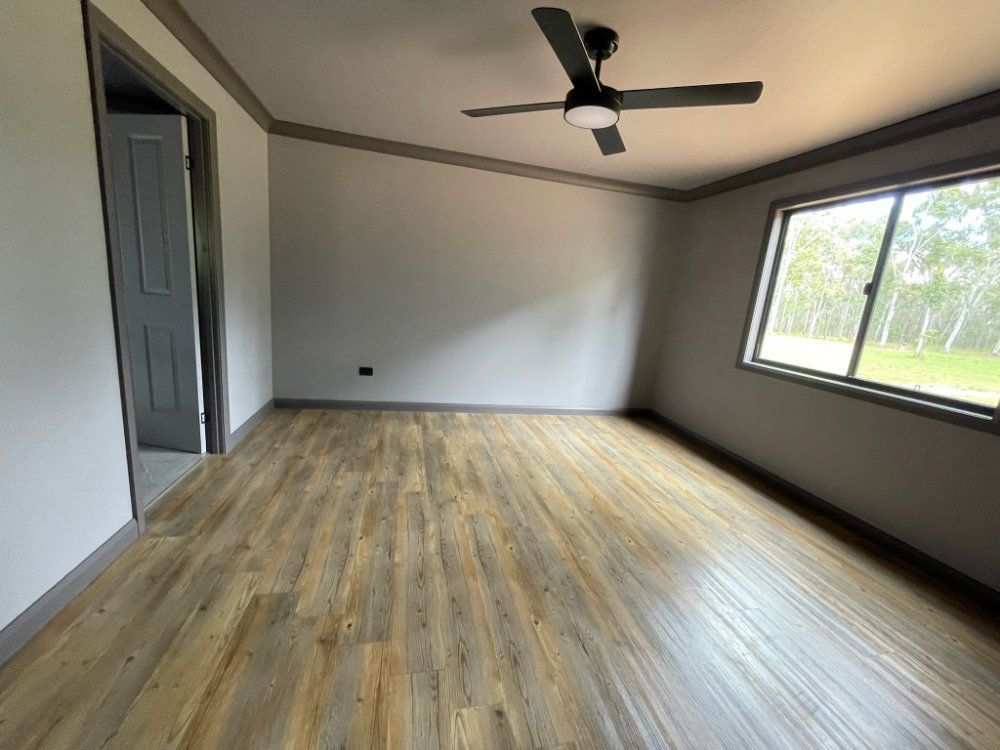 Empty room with wood-look flooring, grey walls, trim. Window, door, and ceiling fan are visible — Steel Homes Australia in Branyan, QLD