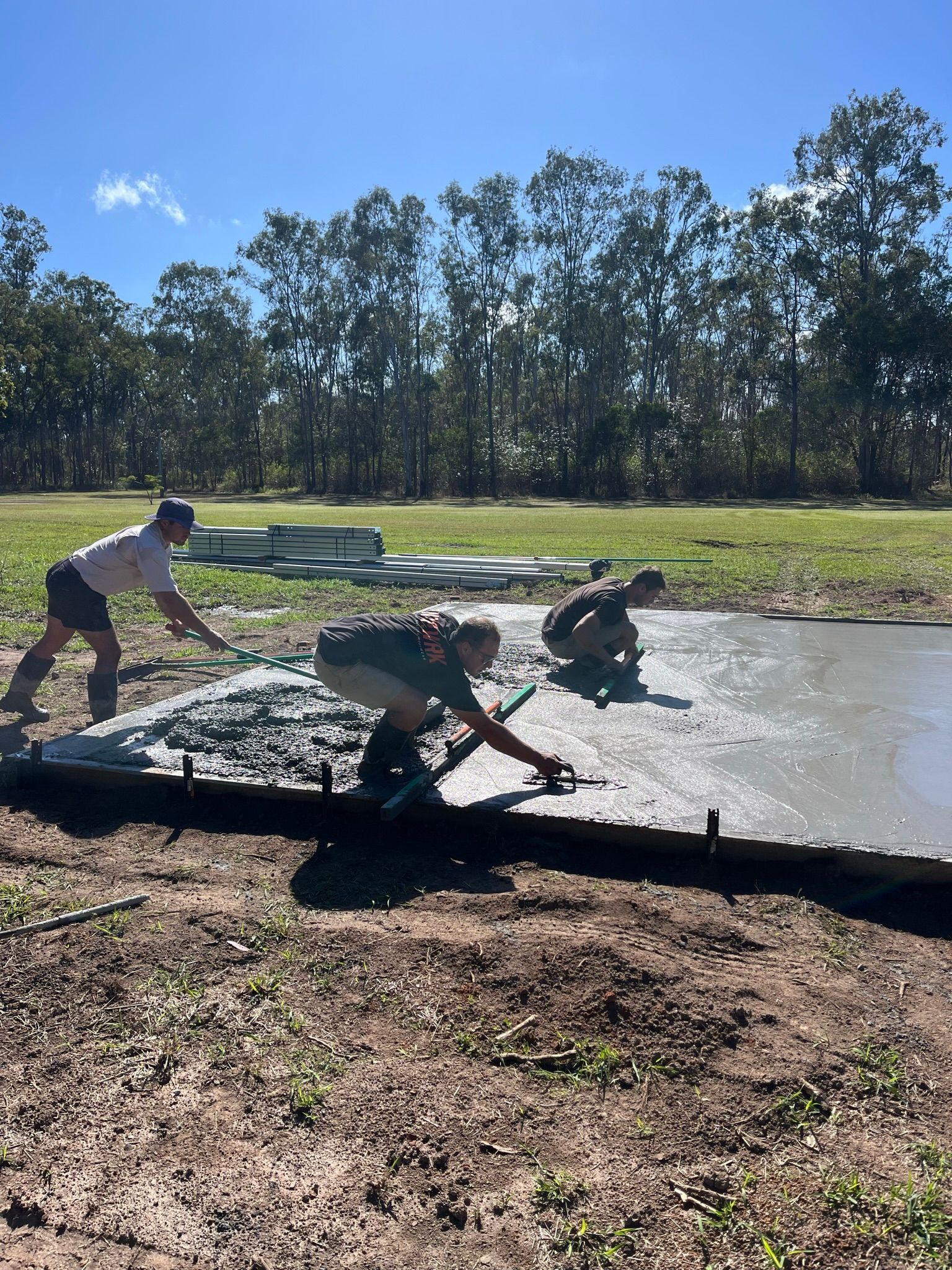 Three people smoothing concrete on a slab outdoors under a sunny sky — Steel Homes Australia in Gin Gin, QLD