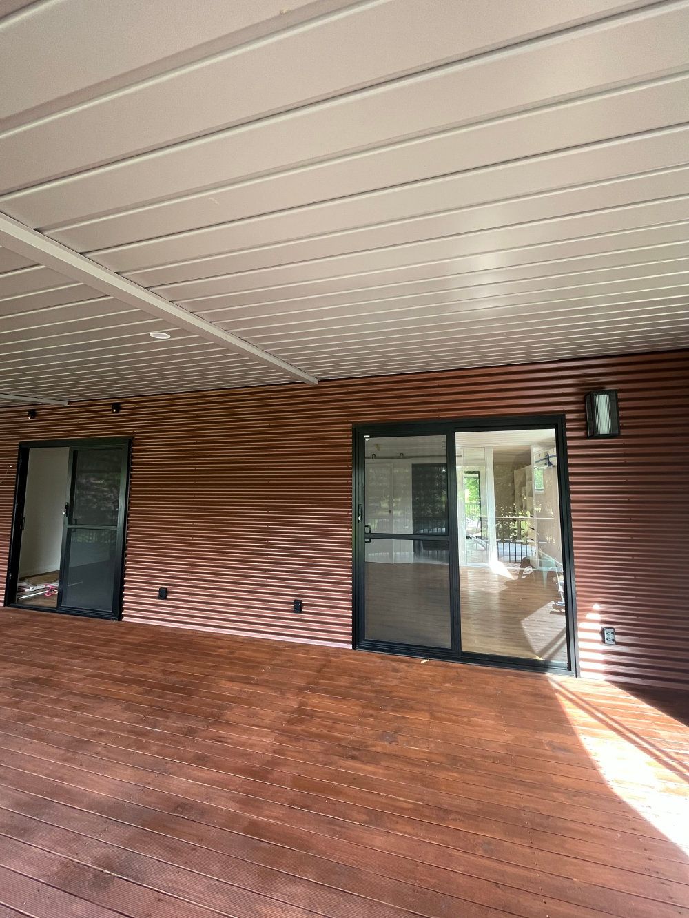 A wooden deck with a brown corrugated wall and black-framed glass doors — Steel Homes Australia in Branyan, QLD