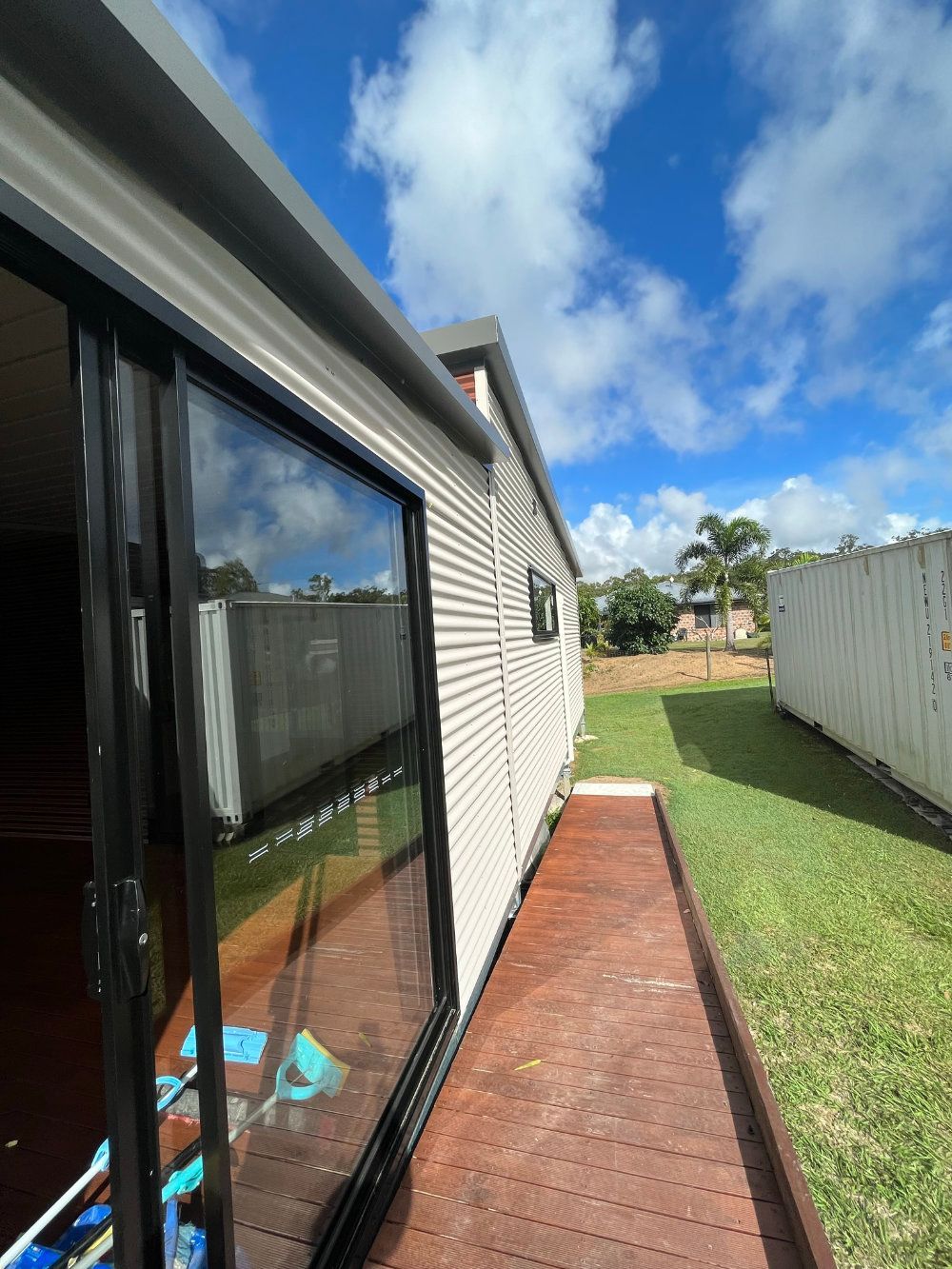 Exterior of a tiny home with black-framed sliding door, wooden walkway, and sunny blue sky — Steel Homes Australia in Branyan, QLD